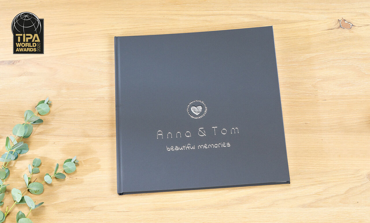 Een CEWE fotoboek met zwarte kaft, gepersonaliseerd met de namen Anna en Tom en de tekst 'beautiful memories'. Het ligt op een lichte houten tafel met een plant op de achtergrond.