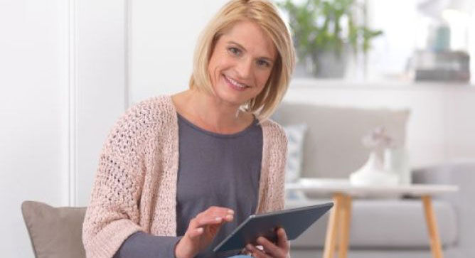 Persoon met een tablet in de hand, zit in een lichte kamer en scrolt blij door inhoud.