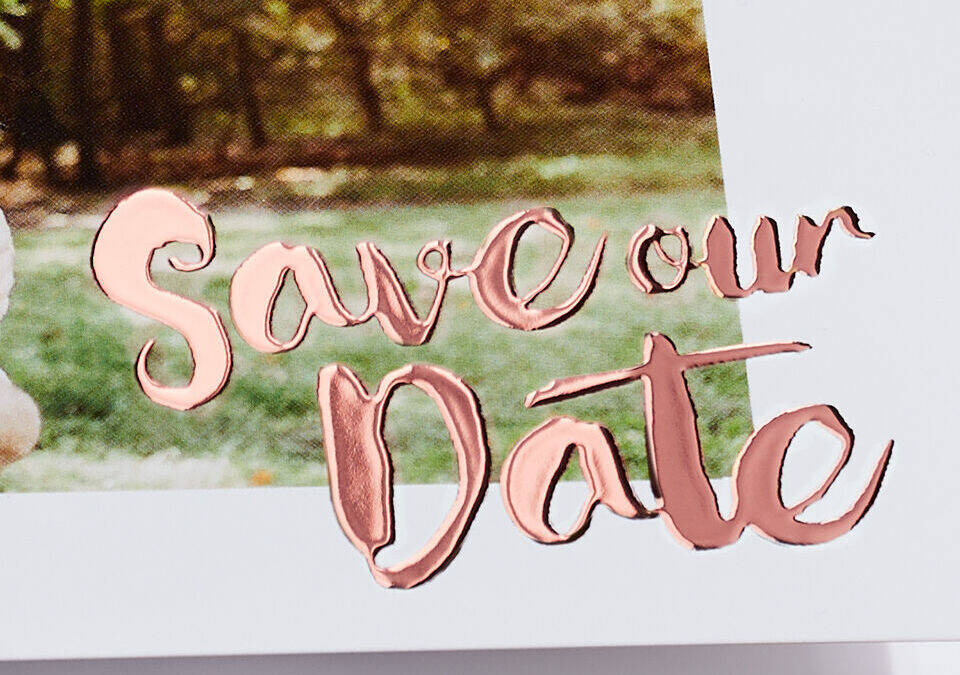 Handgeschreven 'Save our Date' tekst in roségoud op witte achtergrond met groen gras en bomen.