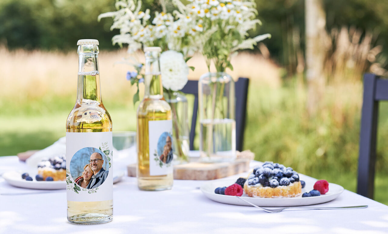 Twee flessen met gepersonaliseerde etiketten op een gedekte tafel, omgeven door bessen en taart.