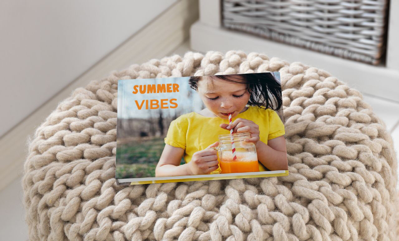 Een vrolijk fotoboek met de titel 'Summer Vibes' op een gezellige poef. Een kind houdt een glas met een oranje drankje vast.
