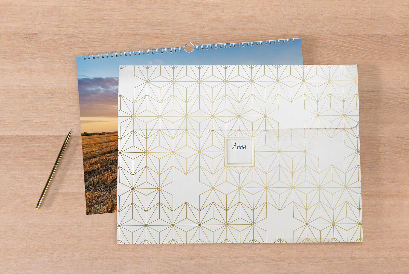 Een liefdevol ontworpen CEWE Fotokalender op een houten tafel, naast een decoratieve envelop met de naam 'Anna' en een gouden pen.