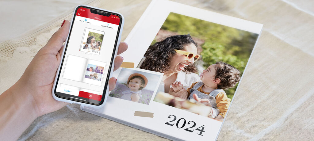 Een hand houdt een smartphone vast met een CEWE fotoboek-preview. Naast een geopend fotoboek met moeder en kind in de zon.