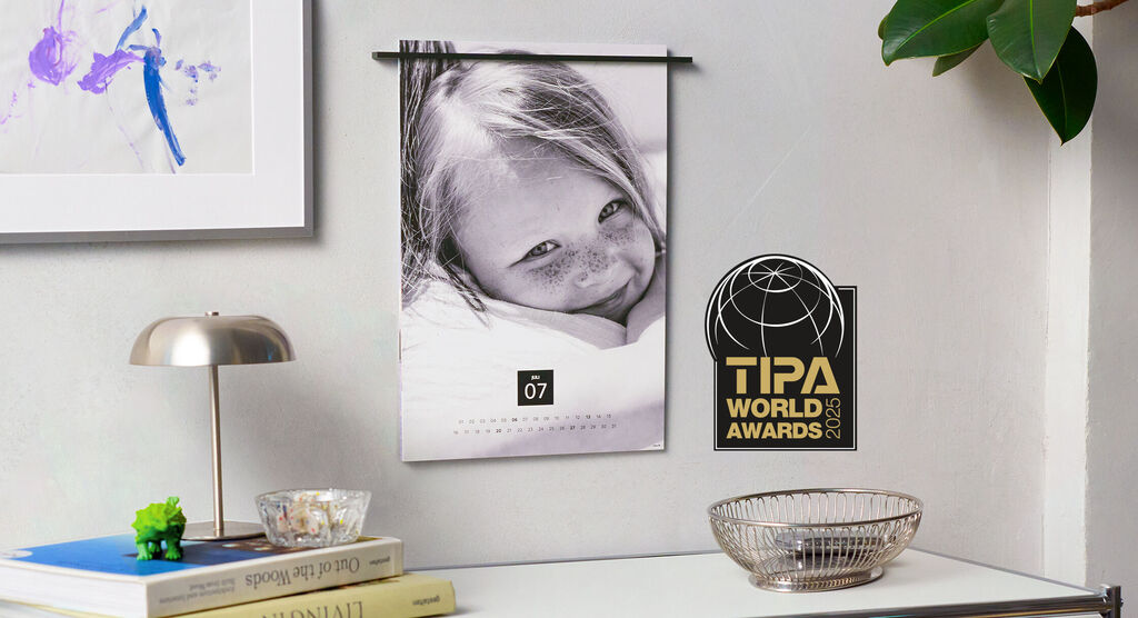 Een mooie kalender hangt aan een muur. Op een tafel liggen een lamp, boeken en een mandje.