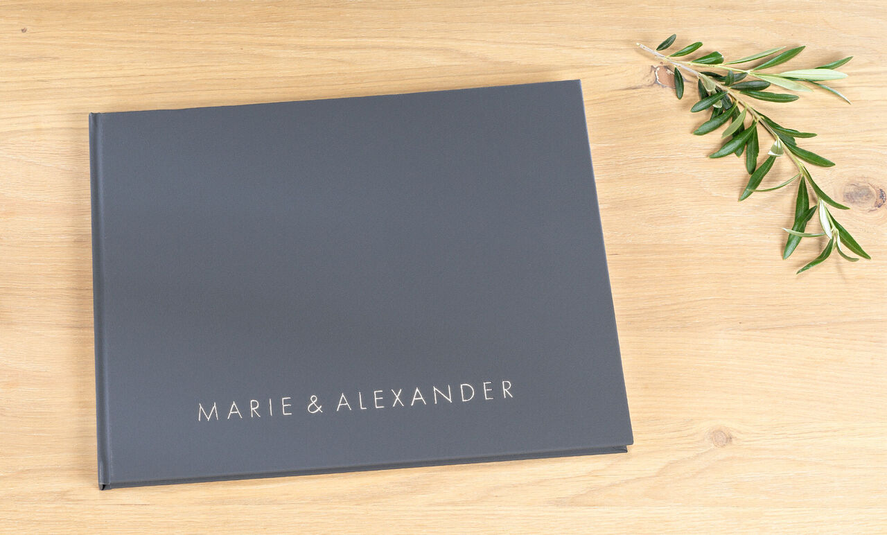 Donker fotoboek met de titel 'Marie & Alexander' op houten ondergrond met olijftak.