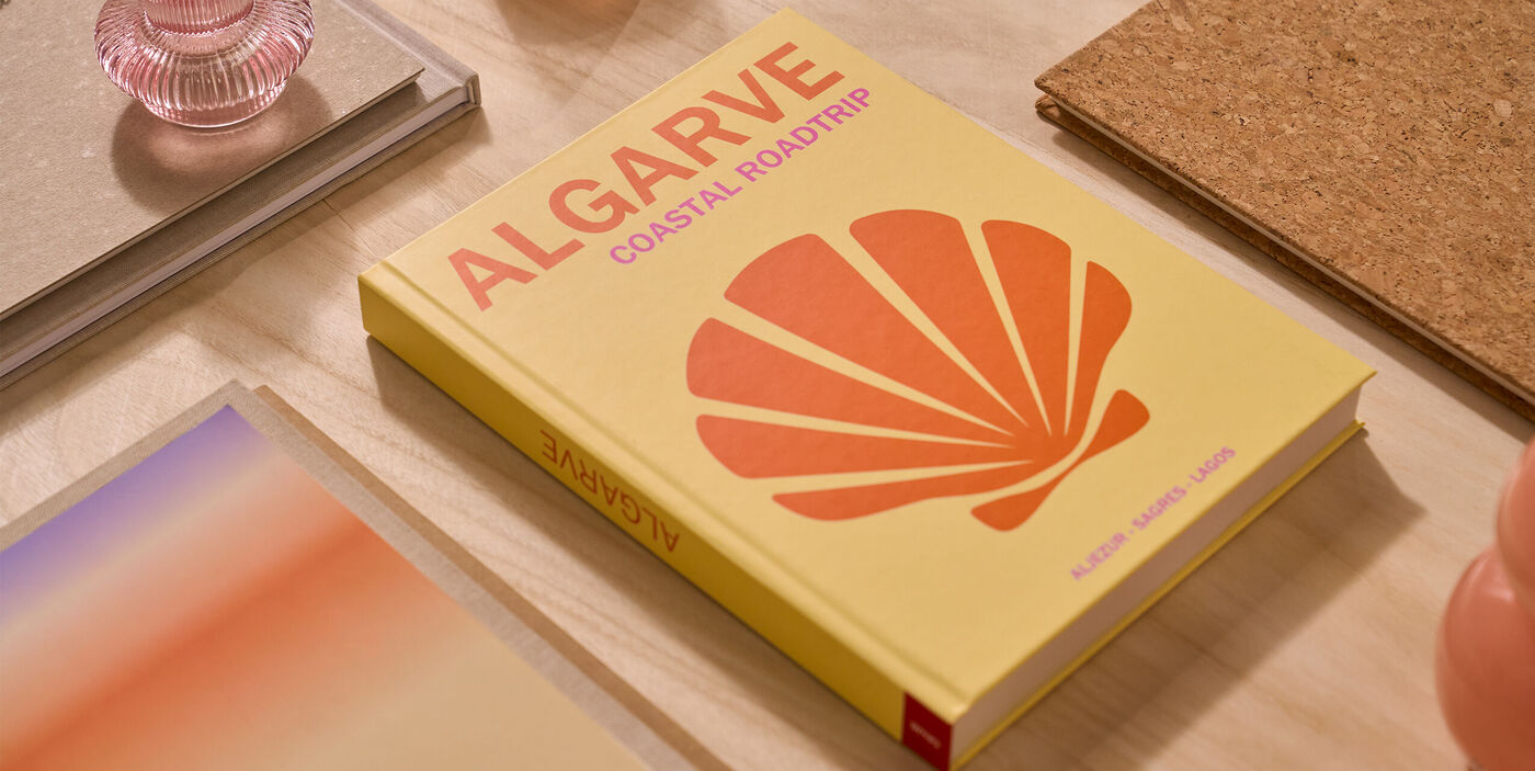 Fotoboek met gele cover en oranje schelp, getiteld “ALGARVE coastal roadtrip”, op een bureau.