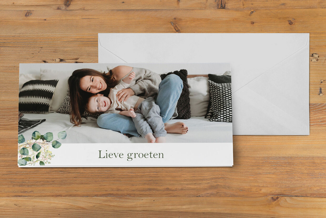 Een moeder en haar kind liggen ontspannen op een sofa en glimlachen gelukkig. Op de achtergrond zijn kussens zichtbaar. Voor hen ligt een kaart met 'Lieve groeten'.