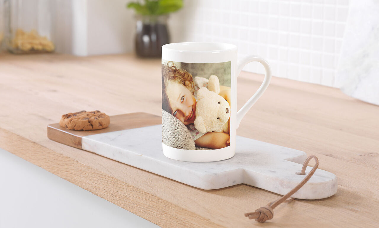 Een kind omarmt een knuffel op een koffiebeker. Op een houten plank ligt een koekje.