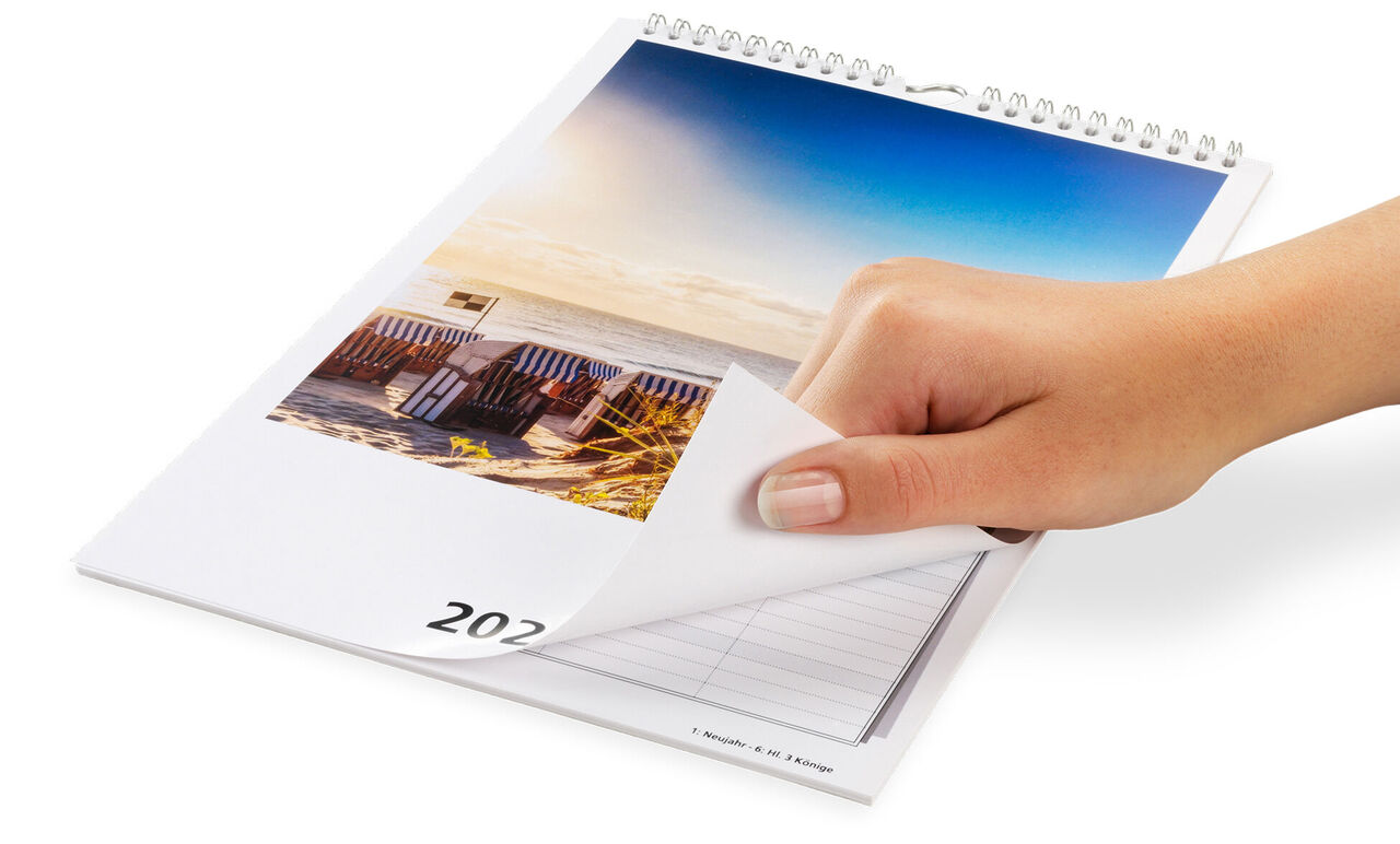 Een hand bladert door een fotokalender met strandmotieven en een mooie hemel.