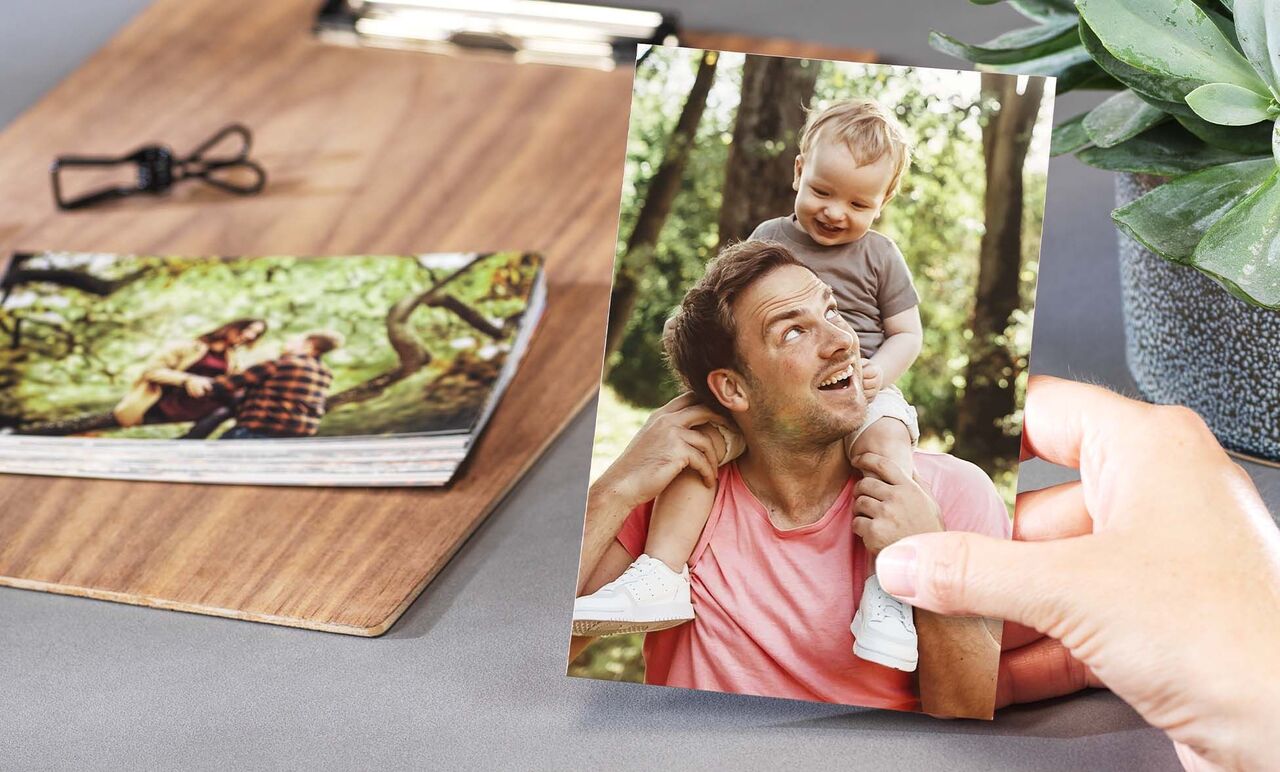 Een hand houdt een foto van een vader met zijn kind op de schouders. Andere foto's liggen op tafel.