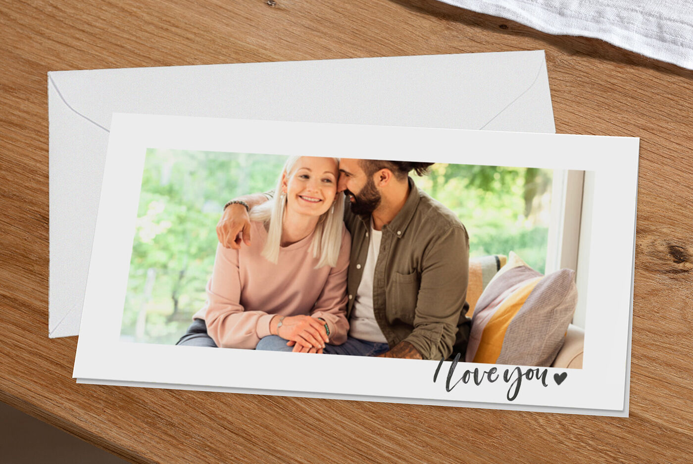 Een vrolijk paar zit samen op een sofa, terwijl ze elkaar liefdevol omarmen en lachen. Op tafel ligt een envelop met 'Love you'.
