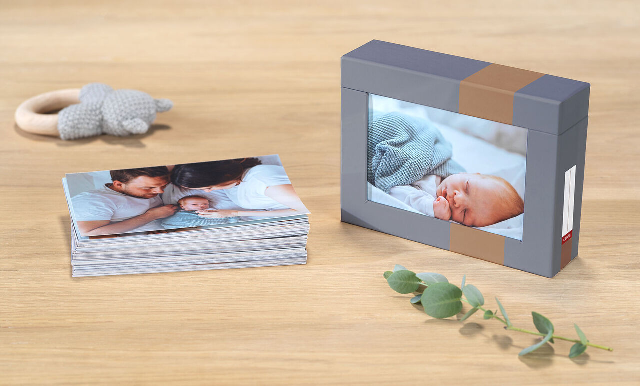 CEWE fotoboek met foto's van een pasgeborene en familie, op een houten tafel naast een doos met babyfoto.