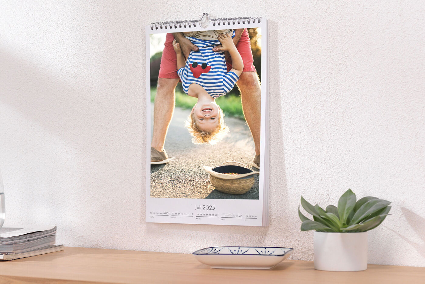 Een kind hangt speels ondersteboven aan een tafel. Een brede lach toont plezier en levensvreugde. Kalender hangt boven houten tafel.