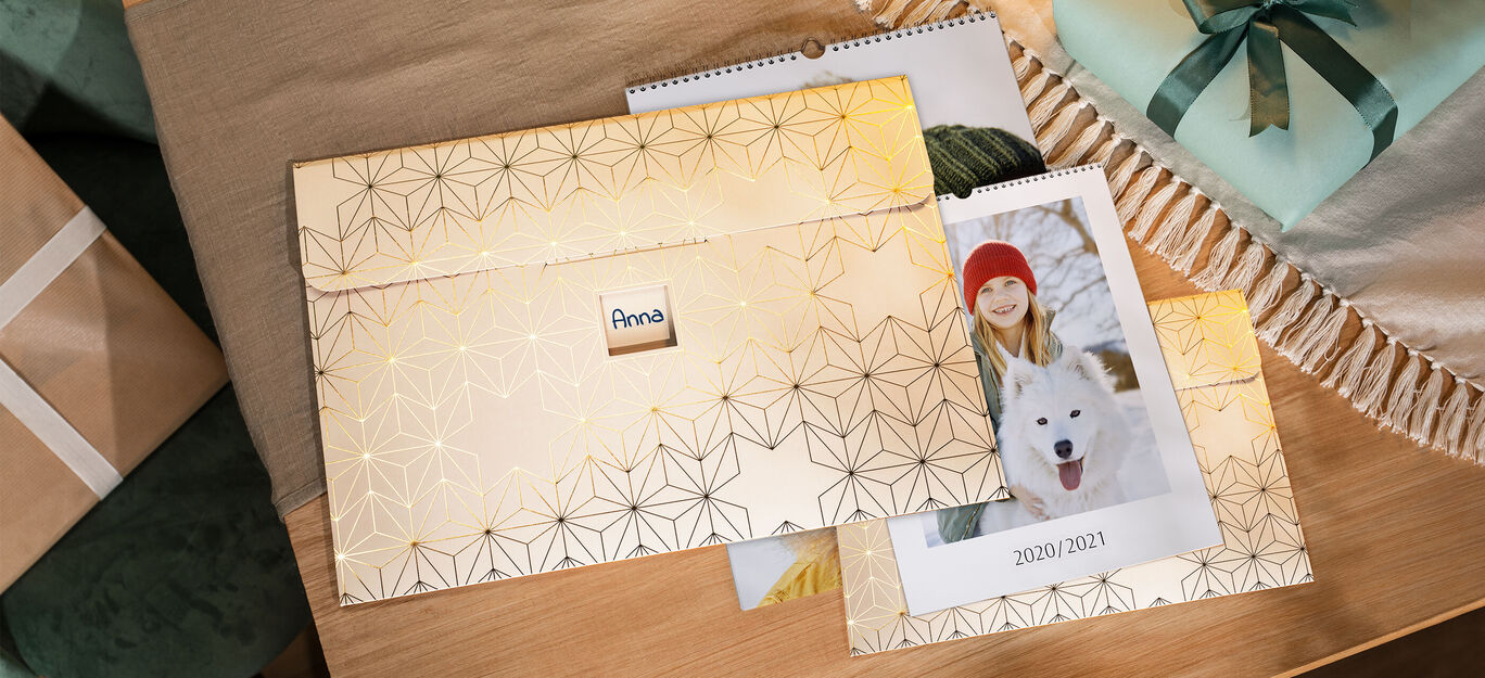 Een envelop van hennepgroetkaarten met gouden decoratie en naam, gelegen op een tafel naast een fotokalender met een vrouw en hond.