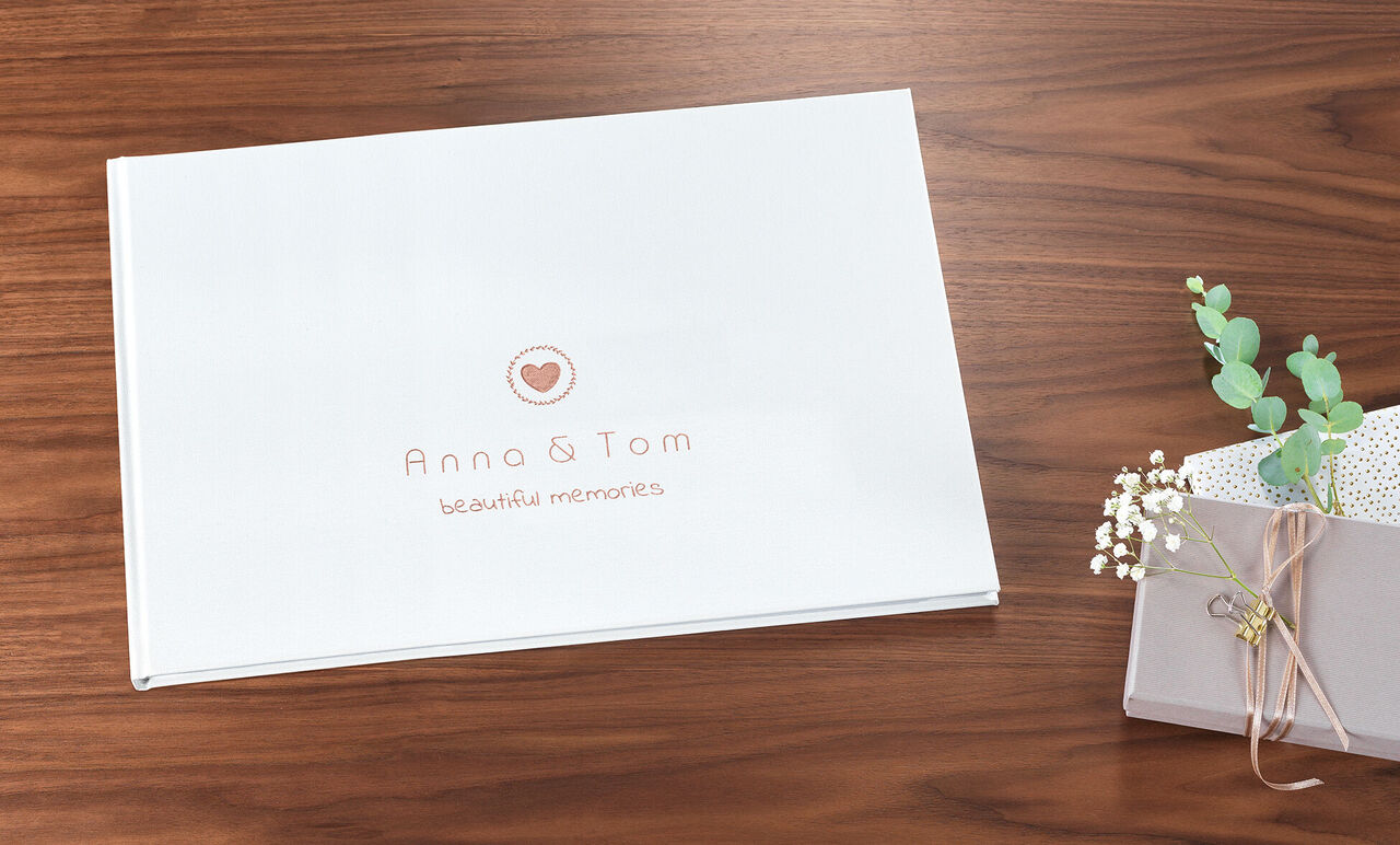 Gepersonaliseerd CEWE FOTBOEK met de titel 'Anna & Tom' op een tafel, omringd door planten en een geschenkdoos.
