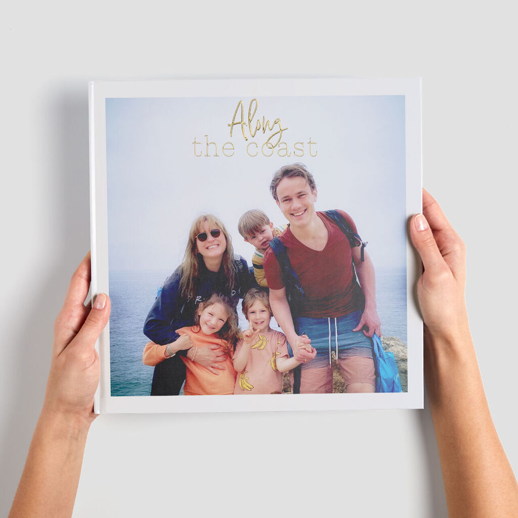 Een persoon houdt een CEWE Fotoboek met de titel 'Alys the coast'. De achtergrond toont de oceaan en lucht.